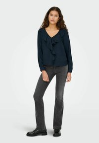 Donkerblauwe lange mouwen top met een V-hals en een geruite voorzijde, gecombineerd met strakke zwarte jeans en zwarte schoenen. Soepele textuur, ontspannen pasvorm.