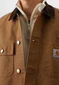 Giacca in tela marrone con colletto in velluto a coste scuro, con tasche sul petto con bottoni e toppa con logo. Indossata sopra uno strato crema con cerniera.