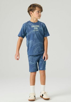 Garçon debout regardant vers la droite, portant un short bleu à imprimé tropical et un T-shirt assorti indiquant «Cooldown Club», avec des chaussettes blanches et des baskets.