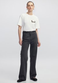 Witte T-shirt met korte mouwen en een paillettenpatroon op de borst, gecombineerd met hoog-getailleerde, wijde, donkergrijze jeans en zwarte versierde slippers.