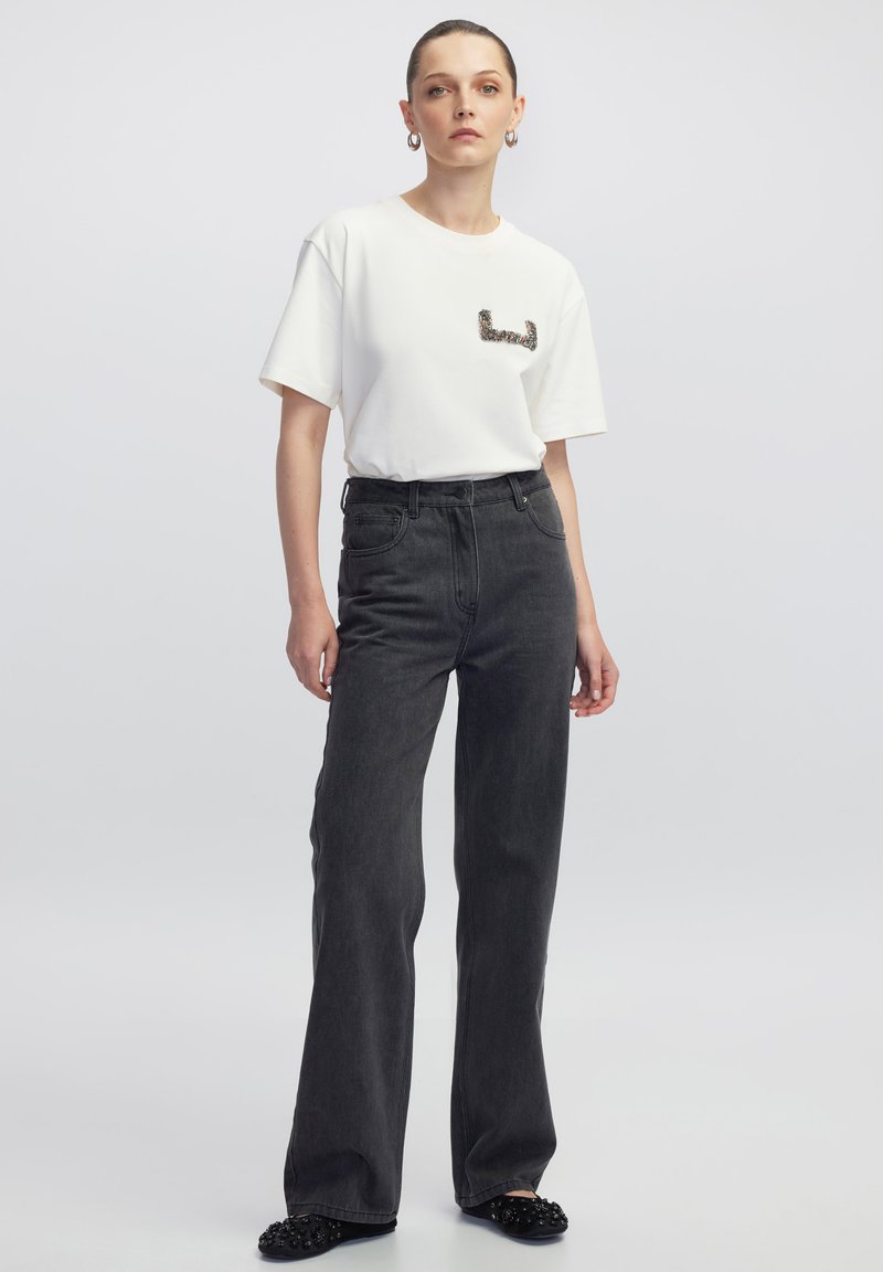 Weißes T-Shirt mit kurzen Ärmeln und einem Paillettenmuster auf der Brust, kombiniert mit hochtaillierten, weiten, dunkelgrauen Jeans und schwarzen verzierten Slippern.