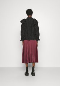 Blusa nera in tessuto testurizzato con colletto arricciato e maniche lunghe, abbinata a una gonna midi plissettata color bordeaux. Stivali neri alla caviglia completano il look.
