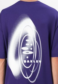 Fioletowa bawełniana koszulka z dużą białą grafiką z koncentrycznymi okręgami oraz wyraźnym napisem "OAKLEY" na dole.