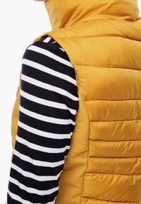 Żółta ocieplana kamizelka o nowoczesnym, puchowym fasonie, zestawiona z granatową i białą w paski bluzą z długim rękawem. Kamizelka ma gładką fakturę.