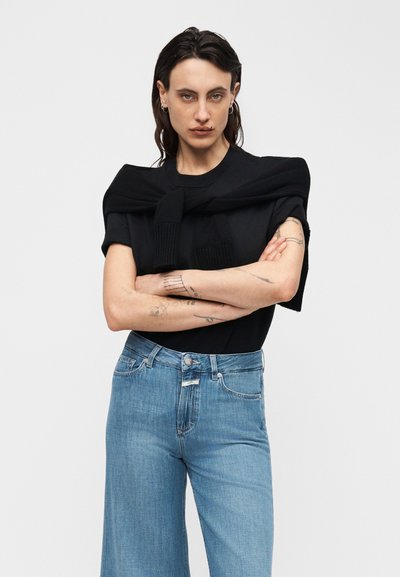 Άτομο με σκούρα μαλλιά και τατουάζ, σταυρώνει τα χέρια, φοράει μαύρο κοντομάνικο μπλουζάκι με πουλόβερ περασμένο και δεμένο στους ώμους, και μπλε τζιν.