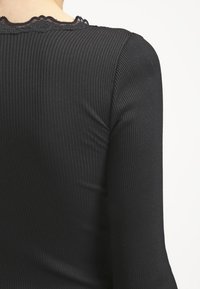 Mustan ribbed pitkähihainen paita, jossa on aallotettu pitsireuna kaula-aukossa. Pehmeä rakenne ja tiukka muotoilu korostavat siluettia.