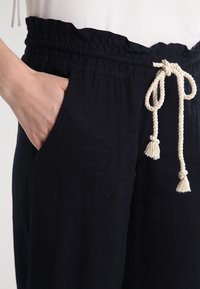 Mörkblå linnbyxor med elastisk midja och krämfärgad dragsko. Har sidofickor och en avslappnad passform som betonar komfort.