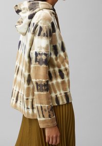 Hoodie a tie-dye marrone e crema con tasca frontale e cappuccio regolabile, dotato di maniche lunghe e vestibilità rilassata, abbinato a una gonna a pieghe.