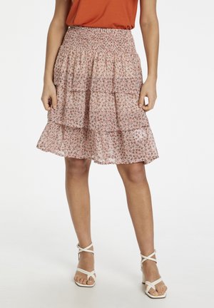 Femme portant une jupe à volants à fleurs roses, un haut orange et des sandales blanches à talons avec des brides, debout devant un fond blanc.