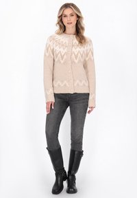 Cardigan bej tricotat cu un model geometric în crem, cu nasturi pe față, decolteu rotund, manșete cu nervuri, purtat peste blugi skinny gri și cizme negre până la genunchi.