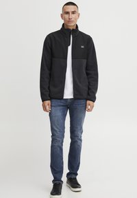 Svart fleecejakke med en glatt svart øvre panel, iført over en hvit skjorte, kombinert med blå jeans og svarte sneakers.