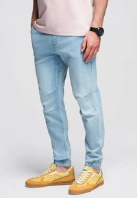 Světlé modré tapered džíny s elastickými manžetami, s bočními švy a nenápadnými nášivkami, doplněné žlutými a zelenými teniskami.