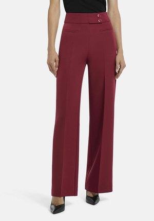 Femme portant un pantalon bordeaux taille haute à jambes larges avec fermeture à double bouton, associé à des talons noirs à bout pointu et un haut noir.