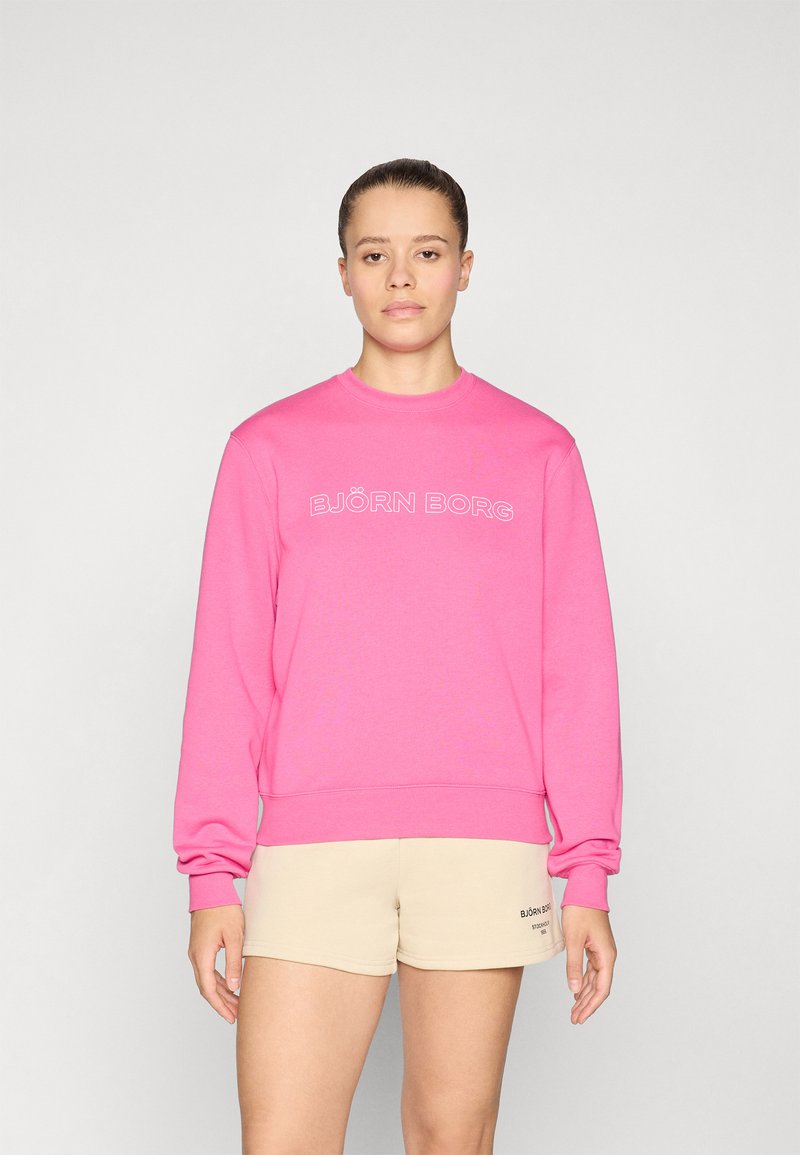 Rosa hettegenser med merket "BJÖRN BORG" i hvite bokstaver, med ribbet mansjetter og nederkant. Brukt med beige shorts.
