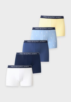 Ung mand står barfodet med armene krydsede, iført lysegule boxershorts med en mørk linning mod en ensfarvet hvid baggrund.