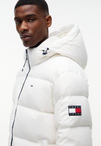 Jeune homme portant une doudoune blanche Tommy Jeans avec capuche, des patchs logo visibles sur la poitrine et la manche, regardant légèrement sur le côté.