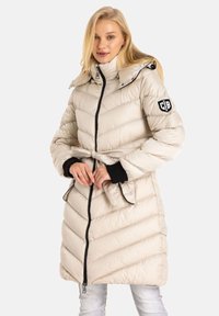 Aufblasbare Jacke in hellem Beige mit einem Gurt in der Taille, Zickzacknähten, hohem Kragen und schwarzen Akzenten an den Ärmeln und dem Reißverschluss. Verfügt über ein Logo-Patch.