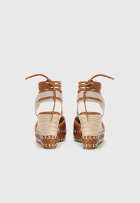 Sandalias de cuña de esparto tejidas en beig con tiras de cuero marrón y lazos en el tobillo, que presentan una suela texturizada y trenzada.