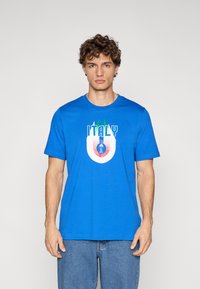 Vīrietis, kurš valkā zilu T-kreklu ar uzrakstu "ITALY" un grafiku zaļā, baltā un sarkanā krāsā, apvienotu ar zilām džinsa biksēm, stāv pret vienkāršu fonu.