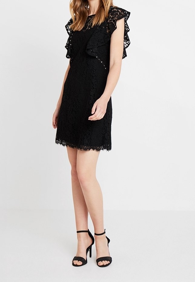 Femme portant une robe noire en dentelle sans manches avec des détails à volants et des talons hauts noirs à bride cheville, debout devant un fond uni.