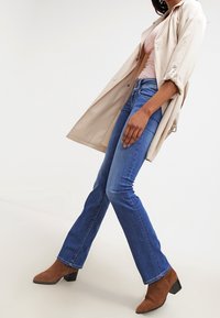 Ljus beige trenchcoat, rosa topp, blå utsvängda denimjeans och bruna ankelstövlar med fyrkantig klack. Slät textur och åtsittande design.