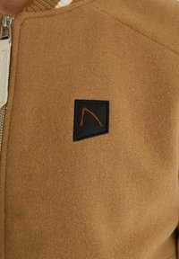 Brązowa kurtka z mieszanki wełny z czarną naszywką z pomarańczowym konturem logo, zamek błyskawiczny i teksturowany, miękki materiał.