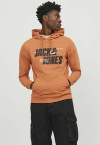 Sudadera con capucha en mezcla de algodón de color naranja quemado, con bolsillo tipo canguro en la parte frontal, capucha con cordón y gran impresión de logo en blanco y negro. Pantalones negros.
