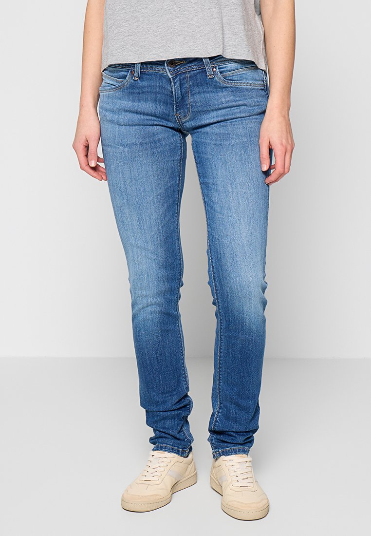 Schmal geschnittene blaue Denim-Jeans mit ausgewaschenen Bereichen, fünf Taschen-Design und mittelhohem Bund, kombiniert mit cremefarbenen Sneakern.