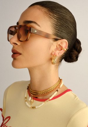 Žena s kosom zalizanom u punđu koja nosi velike sunčane naočale u kornjačevom uzorku, zlatne naušnice u obliku obruča, slojevite ogrlice od zlata i perli te bež majicu.