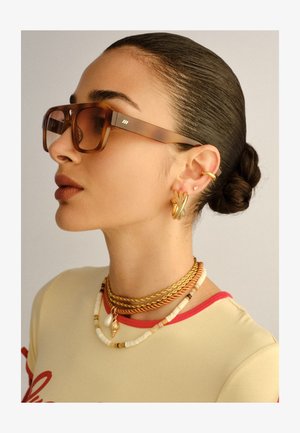 Femme aux cheveux coiffés en arrière en chignon, portant des lunettes de soleil oversize à monture écaille, des boucles d'oreilles créoles en or, des colliers superposés en or et perles, et un haut beige.