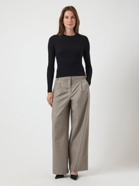 Γυναίκα σε όρθια στάση με μαύρη εφαρμοστή μπλούζα με μακρύ μανίκι, μπεζ παντελόνι σε φαρδιά γραμμή και μαύρα μυτερά παπούτσια, μπροστά σε απλή φόντο.