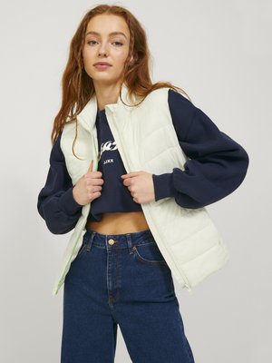 Junge Frau richtet eine ärmellose weiße gesteppte Weste über einem marineblauen, kurz geschnittenen Sweatshirt und hoch taillierten blauen Jeans auf neutralem Hintergrund.