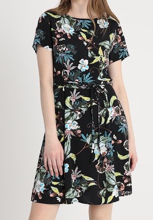 Frau trägt ein kurzärmeliges schwarzes Kleid mit einem Blumenmuster in Blau, Weiß, Grün und Pink, das in der Taille mit einem passenden Gürtel gebunden ist.