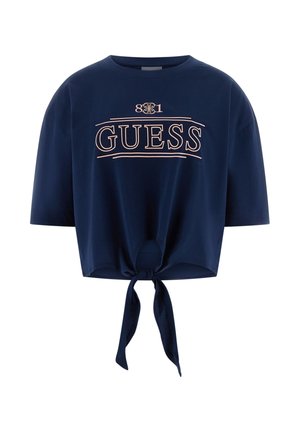 Granatowy krótki t-shirt z krótkimi rękawami, zawiązany na przodzie w supeł oraz napisem "GUESS" w jasnoróżowych literach na wysokości klatki piersiowej.