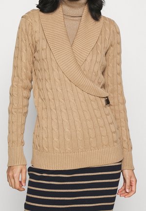 Femme portant un pull cache-cœur beige en maille torsadée avec un grand col côtelé et une jupe marron rayée.