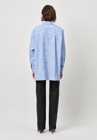 Blue striped shirt with decorative silver accents, featuring a relaxed fit, long sleeves, and a pointed collar, paired with black trousers.