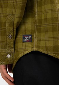 Chemise carrée verte avec un tissu texturé, détails de poignet boutonné et une étiquette avec un graphique "Scotch & Soda" en bas.