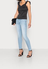 Schwarzes Oberteil mit Polka-Punkt-Muster und Rüsche-Trägern, kombiniert mit hellblauen Skinny-Jeans und schwarzen Blockabsatzsandalen; hält eine schwarze Clutch.