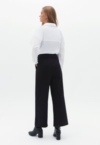 Camicia bianca a maniche lunghe con colletto abbinata a pantaloni neri a vita alta e gamba larga, vista da dietro.