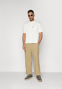 T-shirt bianca a maniche corte con tasca, pantaloni beige larghi e scarpe da ginnastica grigie con accenti neri e beige.