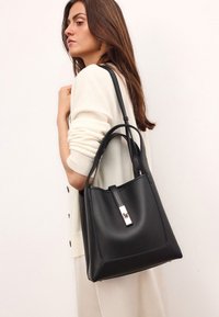 Fekete bőr vödör táska lapos alappal, ezüst hardware-rel és felső pánttal. Simás textúra és varrott részletek az oldalakon.