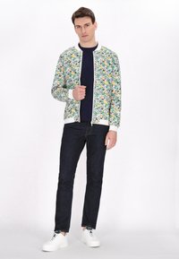 Bloemig bomberjack met een witte basis, versierd met groene bladeren en gele accenten. Gecombineerd met donkere jeans en witte sneakers.