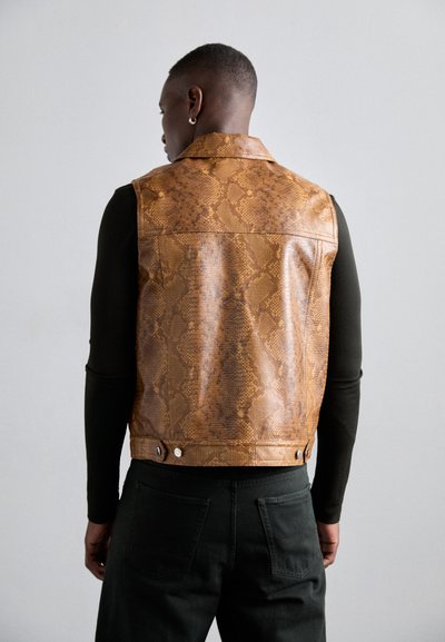 Gilet en simili serpent brun avec un col et fermeture à pression, présentant une texture lisse et un motif détaillé, accompagné de pantalons noirs.
