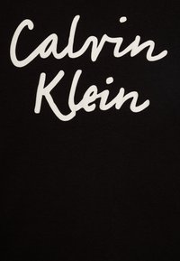 Tela de algodón negro con texto en cursiva blanca prominente que dice "Calvin Klein". El diseño presenta una textura suave con detalles mínimos.