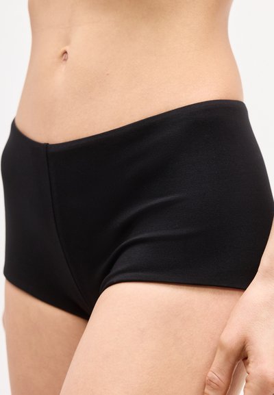 Γυναικείο κορμί που φορά μαύρα άνευ ραφών εσώρουχα, εμφανίζοντας το κάτω μέρος της κοιλιάς, τους γοφούς και μέρος του δεξιού χεριού που ακουμπά στο πλάι.