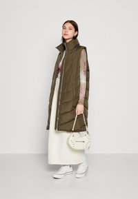 Gilet rembourré vert olive avec col montant, fermeture éclair devant et poches latérales, associé à un haut floral léger, une jupe blanche et des baskets blanches.