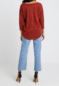 Haut rouge texturé, ample, avec un ourlet arrondi et des manches trois-quarts, porté avec un jean court bleu clair et des sandales à talons noirs.