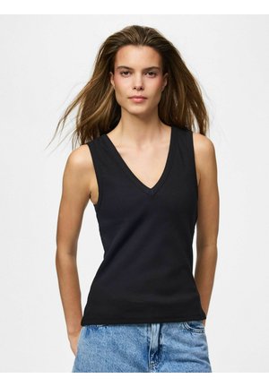 Žena s dlhými hnedými vlasmi, oblečená v čiernom bezrukávovom top s výstrihom do V a modrých džínsoch, čelom k prednej strane na jednoliatom pozadí.