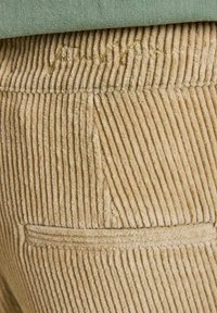 Pantalons en velours côtelé beige clair, avec une texture de côtes verticales. Comprend une petite poche arrière et un détail de couture doré discret.