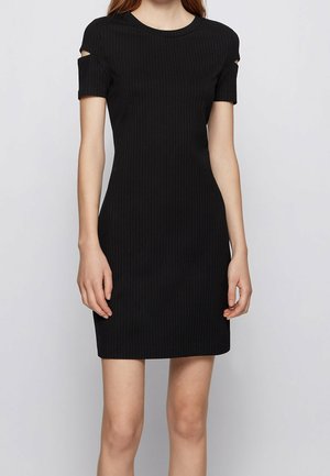 Femme portant une courte robe noire côtelée avec des détails découpés sur les manches courtes, debout devant un fond uni gris clair.